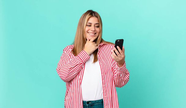curvy pretty blonde woman smiling with a happy, confident expression with hand on chin and holding a smart phone - Фото, зображення