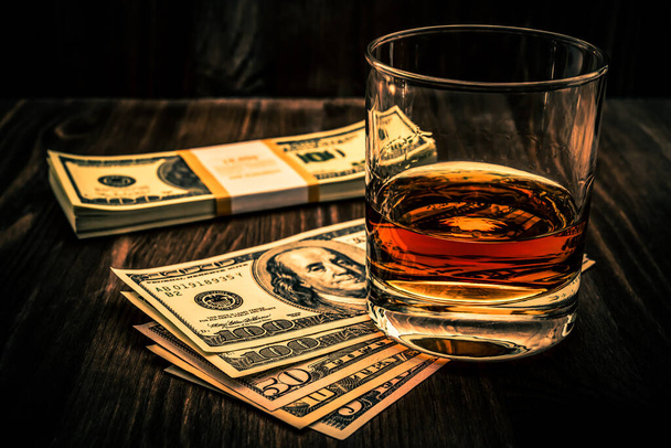 Glass of whiskey with pack of dollars on a wooden table. Angle v - Photo, image