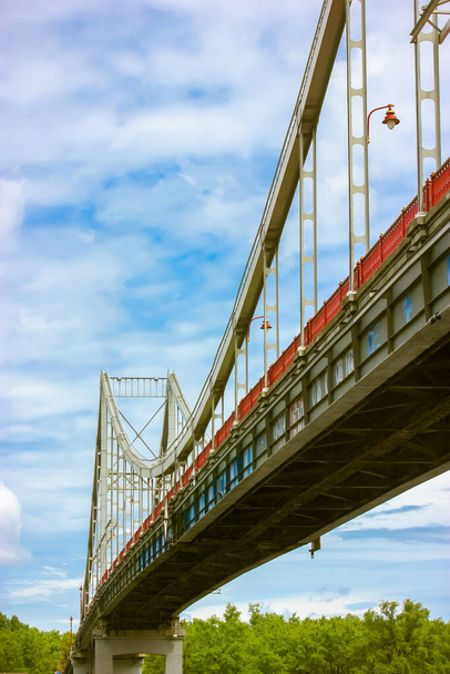 Eine moderne eiserne Schrägseilbrücke mit Tragkonstruktion und Laterne vor blauem wolkenverhangenem Himmel an Sommertagen. Blick von unten auf eine hohe rote Brücke in Kiew, Ukraine. Urbane Architektur. - Foto, Bild