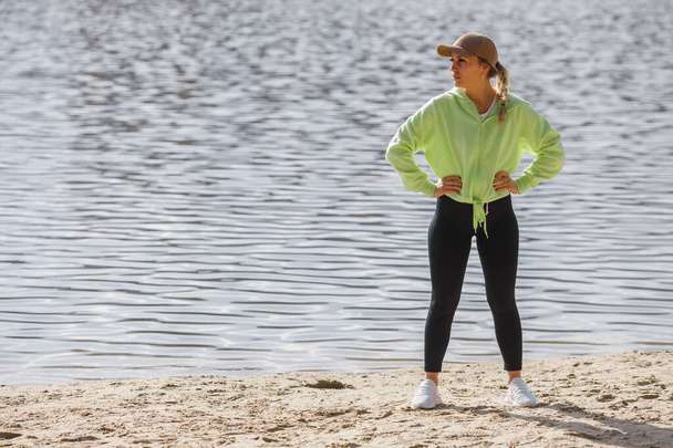 Itsetuhoinen naisjuoksija trendikkäissä urheiluvaatteissa, jotka poseeraavat joenrannalla, venyttävät jalkoja, lämmittävät lihaksia ennen juoksemista ulkona aikaisin aamulla.. - Valokuva, kuva