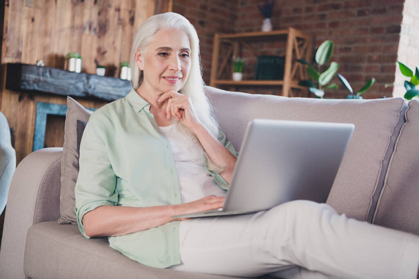 Фото милой очаровательной пенсионерки носить зеленую рубашку сидя диван печатая современное устройство улыбаясь в помещении дома квартиру - Фото, изображение