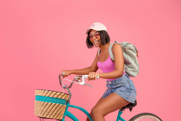 Actividades de verano al aire libre. Longitud completa de la mujer negra despreocupada montar en bicicleta, divertirse, hacer deportes sobre fondo rosa - Foto, imagen