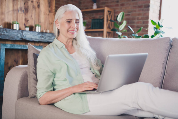 Portrait of attractive focused cheerful granny sitting on sofa reading book surfing web using laptop at home brick loft interior house indoor - Zdjęcie, obraz