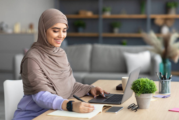 Muzułmanka siedząca za biurkiem, używająca komputera do pisania w notatniku - Zdjęcie, obraz