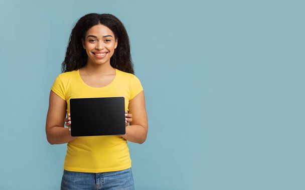 Super stránka. Šťastná černá dáma ukazuje tablet s prázdnou obrazovkou, stojící nad modrým studiem pozadí s volným místem. - Fotografie, Obrázek