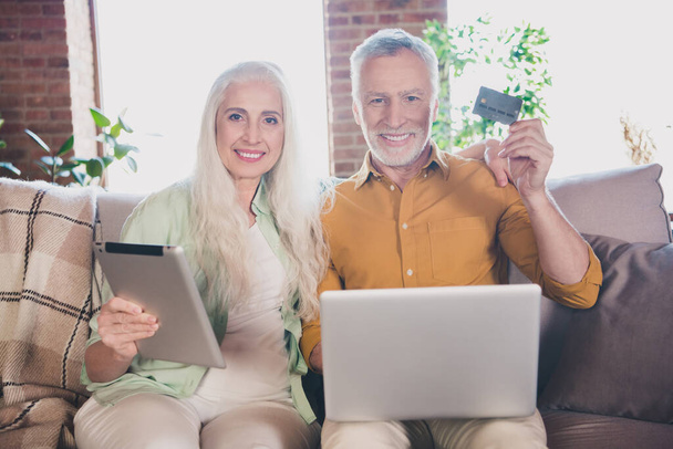 Photo of grey haired retired pensioner friends wife husband couple sit couch hold credit card tablet laptop indoors inside house home - Fotoğraf, Görsel