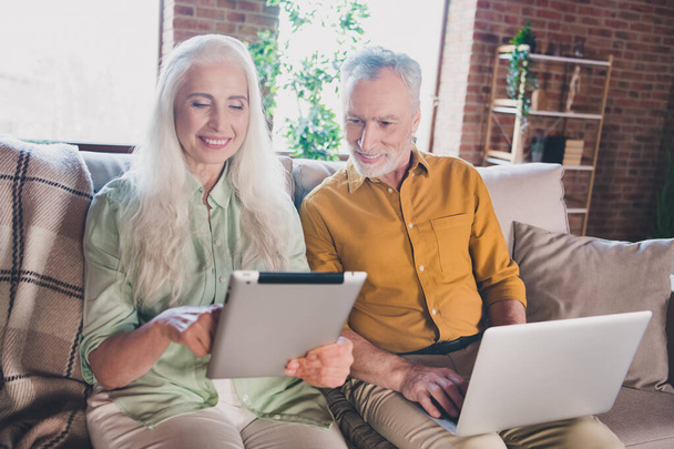 Photo of charming happy old grey haired people smile sit sofa use tablet laptop indoors inside house apartment - Foto, Imagem