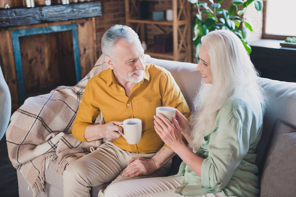 Photo of positive charming man and woman old people good mood sit sofa drink tea indoors inside house home - Foto, afbeelding