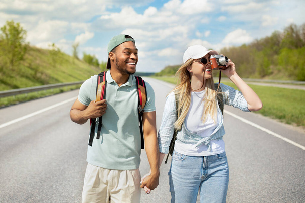 Joyeux jeune couple multiracial faisant du stop ensemble, se tenant la main, prenant des photos à la caméra, marchant le long de la route - Photo, image