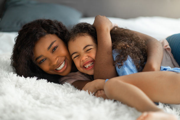 Rodzicielska miłość dziecka. Szczęśliwy młody czarny kobieta obejmując jej córka na łóżku, słodkie dziewczyny śmiejąc się i ciesząc się przytulić - Zdjęcie, obraz