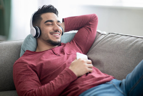 Tipo árabe positivo disfrutando de la música, utilizando el teléfono móvil y auriculares - Foto, imagen