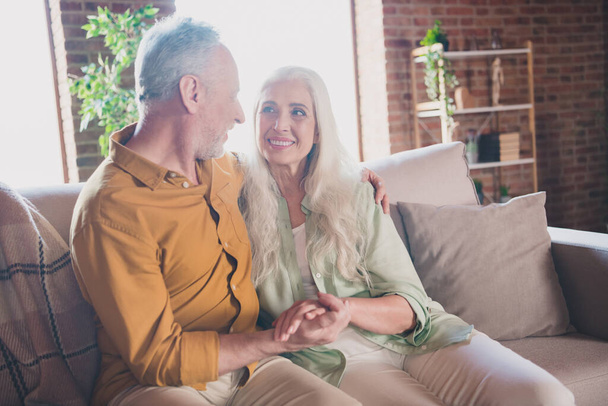 Portrait of attractive cheerful affectionate grey-haired couple sitting on sofa holding hands at home loft interior house flat indoor - Foto, Imagem