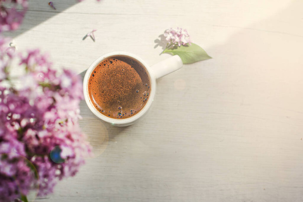 Un mazzo di fiori di lilla sulla finestra e caffè in una tazza bianca sotto i raggi del sole. Un bouquet di lillà e una tazza di caffè o tè.  - Foto, immagini