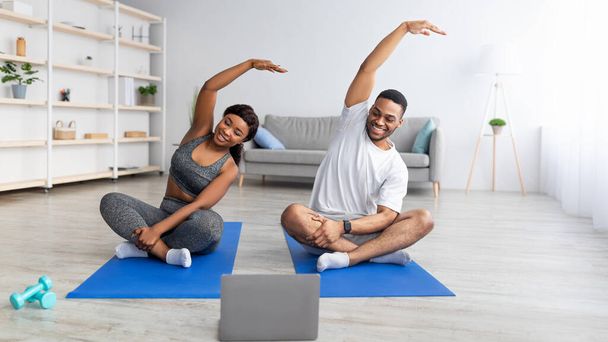Sportowa czarna para siedząca w pozycji lotosu na matach fitness, rozciągająca ramiona klasą jogi online w domu - Zdjęcie, obraz