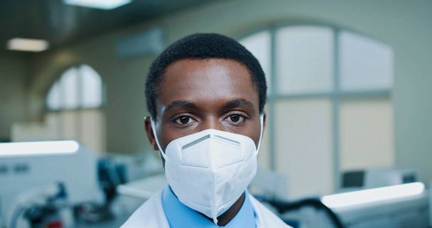 Close up shot of handsome African American young male doctor with protective mask on face looking at camera at workplace in clinic lab. Male doctor in laboratory, hospital, coronavirus pandemic - 写真・画像