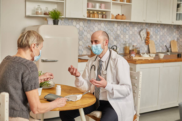 Man doctor wearing a protective mask advises an elderly woman wearing a protective mask at her home. - Fotó, kép