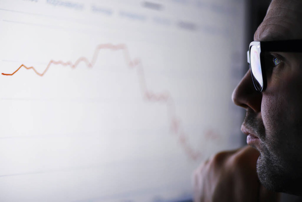 Un hombre de negocios está mirando un gráfico en un monitor. Un corredor de bolsa evalúa las tendencias del mercado de valores. Un hombre con gafas frente a una curva de la dinámica de la economía. - Foto, imagen