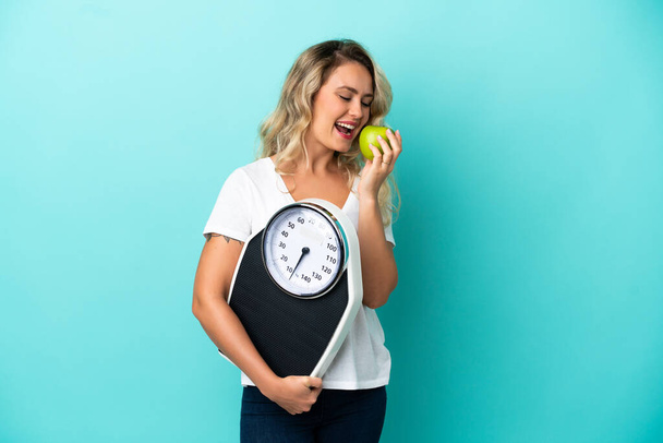 Young Brazilian woman isolated on blue background with weighing machine and with an apple - Fotografie, Obrázek