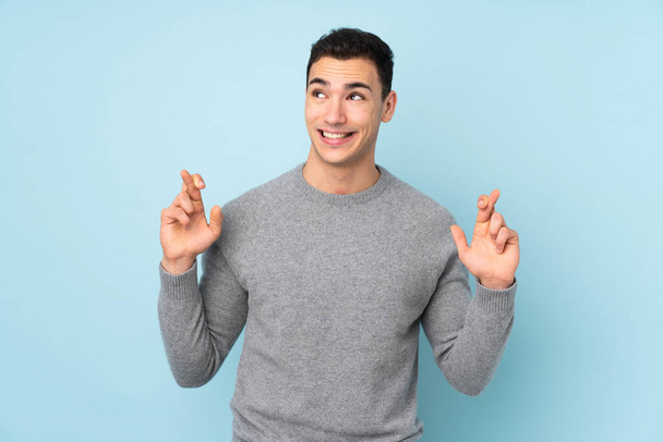 Young caucasian handsome man isolated on blue background with fingers crossing - Photo, Image