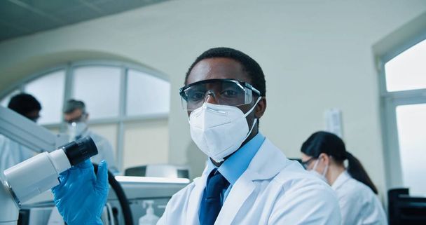 Close up portrait of young handsome African American male medical scientist work in clinic lab on modern equipment on virus cure. Healthcare specialist in mask and goggles looking through microscope - 写真・画像