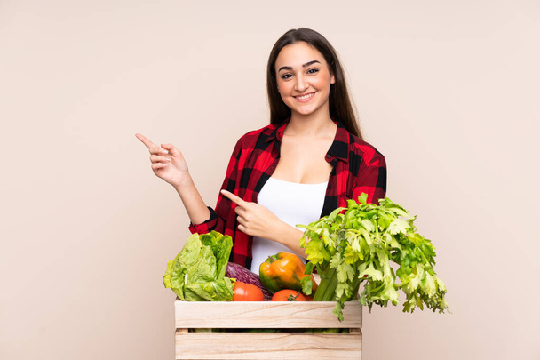 Landwirt mit frisch gepflücktem Gemüse in einer Box isoliert auf beigem Hintergrund zeigt mit dem Finger zur Seite - Foto, Bild