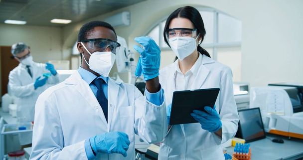 Close up portrait of African American young male speak with Caucasian female medical scientist in mask standing in clinic lab and typing on tablet discussing covid cure in glassware, medicine concept - Фото, изображение