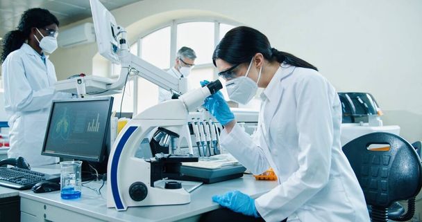 Side view of young Caucasian female medical scientist in mask and gloves sitting at laboratory using modern technology looking in microscope conducting experiment. Lab assistant making analysis - Foto, afbeelding