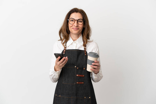 Restaurante de mediana edad camarera mujer aislada sobre fondo blanco sosteniendo café para llevar y un móvil - Foto, Imagen