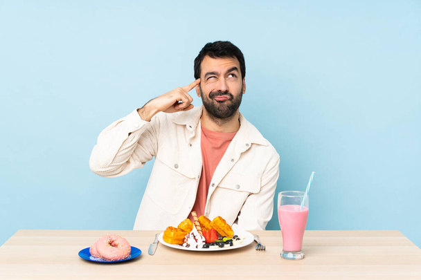 Человек за столом, завтракающий вафлями и молочным коктейлем, делая жест безумия пальцем по голове - Фото, изображение