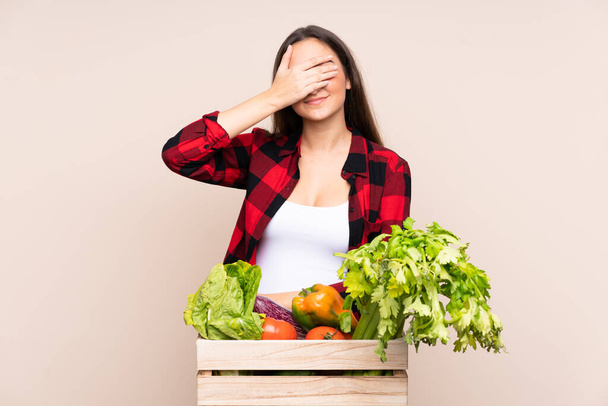 Rolnik ze świeżo zebranymi warzywami w pudełku odizolowanym na beżowym tle pokrywającym oczy dłońmi - Zdjęcie, obraz