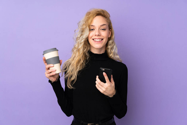 Νεαρή ξανθιά γυναίκα απομονωμένη σε μωβ φόντο κρατώντας καφέ για να πάρει μακριά και ένα κινητό - Φωτογραφία, εικόνα