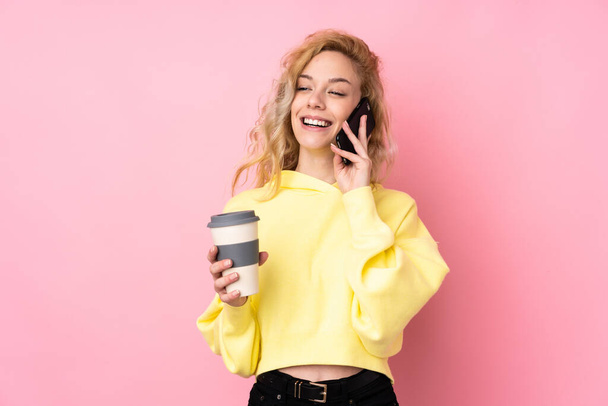 Young blonde woman wearing a sweatshirt isolated on pink background holding coffee to take away and a mobile - Foto, immagini