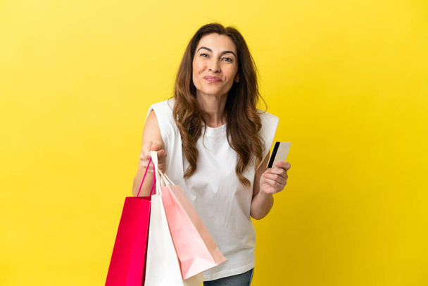 Mujer caucásica de mediana edad aislada sobre fondo amarillo sosteniendo bolsas de compras y una tarjeta de crédito - Foto, Imagen