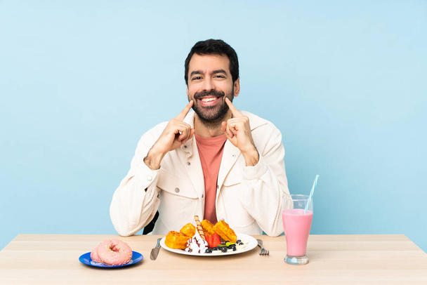 Ο άνθρωπος σε ένα τραπέζι τρώει βάφλες και ένα μιλκσέικ χαμογελώντας με μια χαρούμενη και ευχάριστη έκφραση - Φωτογραφία, εικόνα
