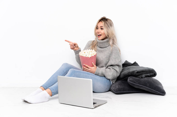 Teenager blonde girl eating popcorn while watching a movie on the laptop pointing finger to the side and presenting a product - Photo, Image