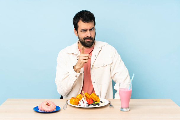 Ο άνθρωπος σε ένα τραπέζι τρώει βάφλες και ένα μιλκσέικ απογοητευμένο και δείχνει προς τα εμπρός - Φωτογραφία, εικόνα