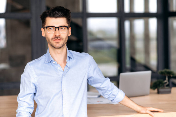 Portrait of a smart handsome confident caucasian businessman or freelancer, with glasses, stands near his work desk in the office, in looks directly into the camera, resting his hand on the table - Photo, Image