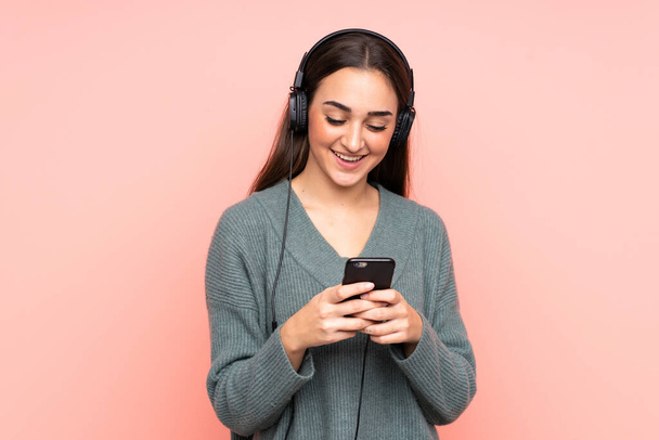 Young caucasian woman isolated on pink background listening music and looking to mobile - Foto, Bild