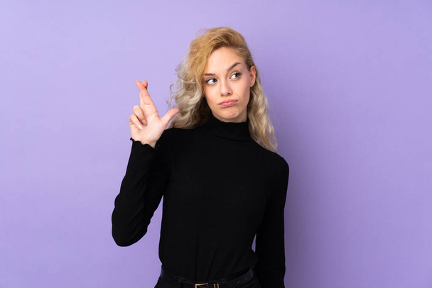 Jeune femme blonde isolée sur fond violet avec les doigts croisés et souhaitant le meilleur - Photo, image