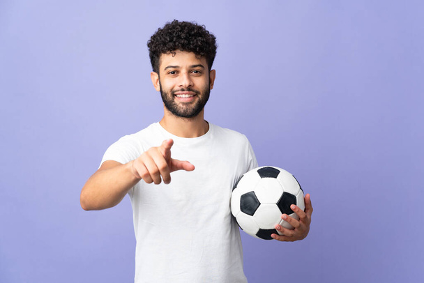 Joven hombre marroquí aislado sobre fondo púrpura con pelota de fútbol y apuntando hacia el frente - Foto, imagen