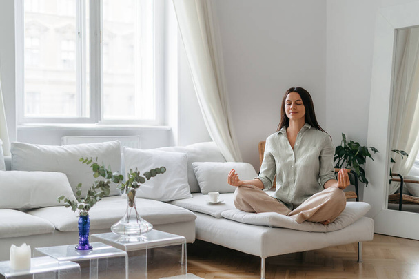 Giovane bella donna bruna medita mentre si siede in posa yoga loto seduto sul divano bianco in soggiorno - Foto, immagini