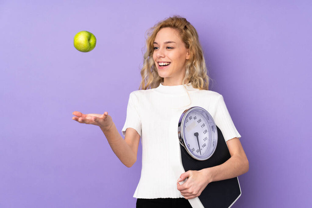 Junge blonde Frau isoliert auf lila Hintergrund mit Wiegemaschine und mit einem Apfel - Foto, Bild