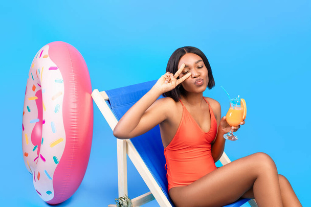 Flirty black lady with tropical cocktail relaxing in lounge chair, pursing her lips for kiss, making peace gesture - Foto, Imagen