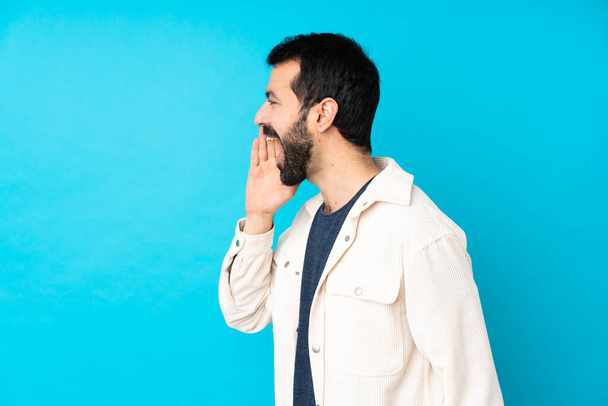 Jovem homem bonito com jaqueta de veludo branco sobre fundo azul isolado gritando com a boca bem aberta para o lateral - Foto, Imagem