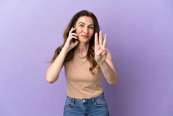 Середньовічна біла жінка використовує мобільний телефон ізольовано на фіолетовому фоні щаслива і рахує трьох пальцями
 - Фото, зображення