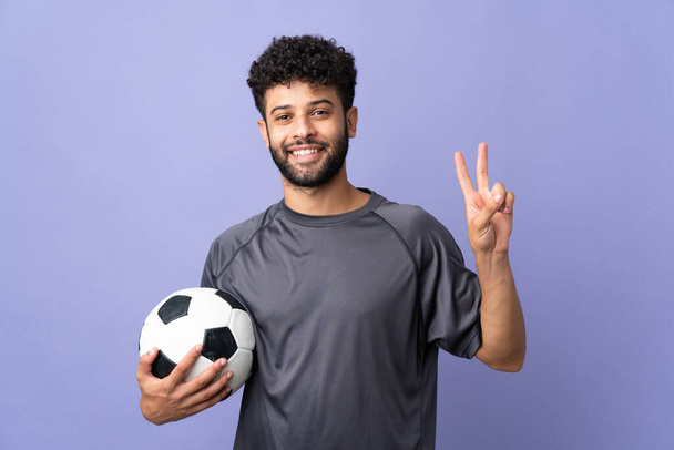 Bonito marroquino jovem jogador de futebol homem sobre isolado no fundo roxo sorrindo e mostrando sinal de vitória - Foto, Imagem