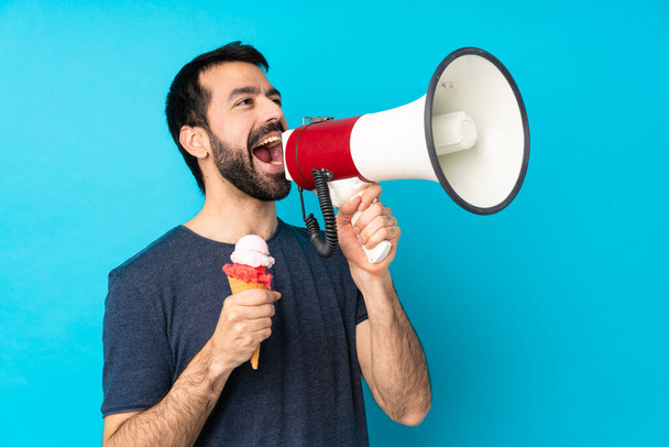 Молодой человек с мороженым из корнета на изолированном синем фоне кричит через мегафон - Фото, изображение