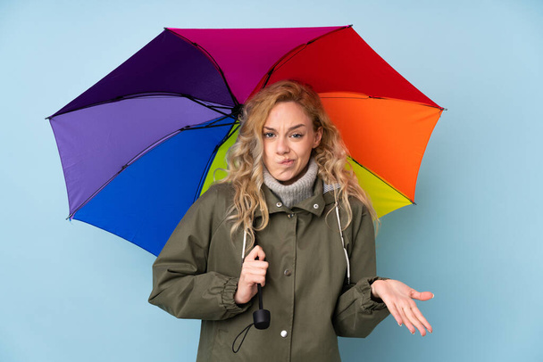 Jovem loira segurando um guarda-chuva isolado no fundo azul fazendo gestos de dúvidas ao levantar os ombros - Foto, Imagem