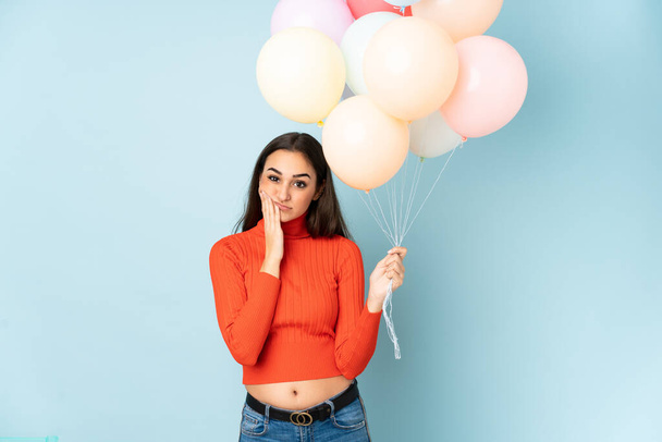 Młoda kobieta łapie wiele balonów odizolowanych na niebieskim tle nieszczęśliwa i sfrustrowana - Zdjęcie, obraz