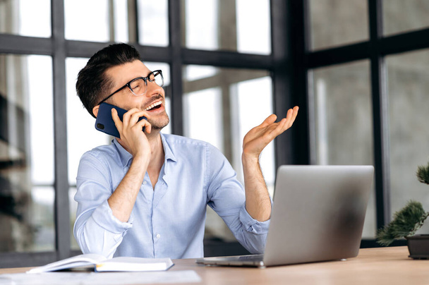 Зв'язок з мобільним телефоном. Дружній радісний кавказький бізнесмен сидить за столом в офісі, емоційно спілкується по смартфону з друзями або колегами, жести рукою, дивиться вбік, посміхається
 - Фото, зображення